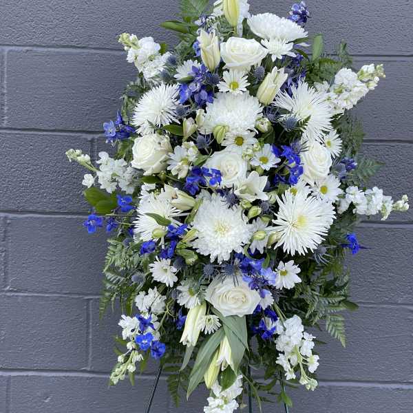 White and blue funeral spray with roses, lilies, and daisies on a stand
