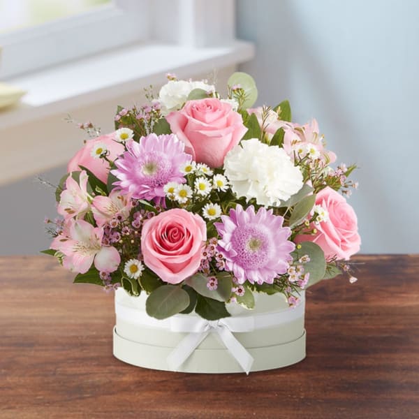 Pink roses and mixed blooms arranged in a round white box with a ribbon