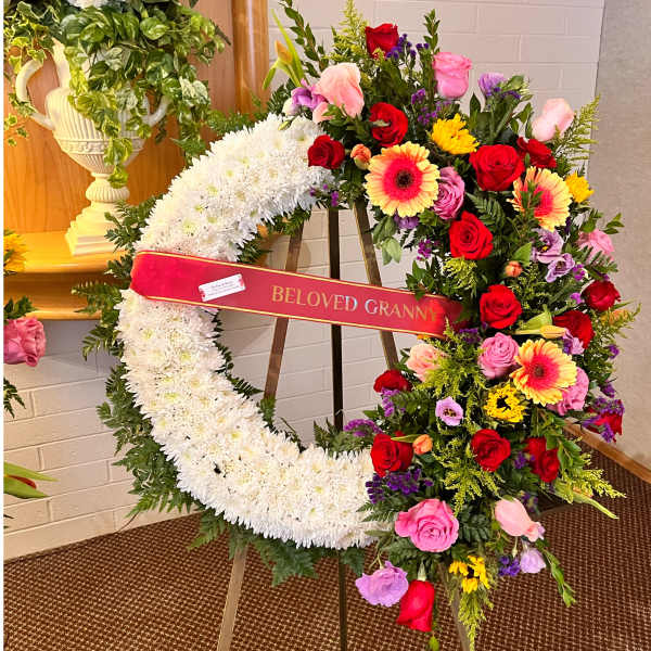 Standing white sympathy wreath with bright mixed flowers and a 'Beloved Granny' ribbon on an easel