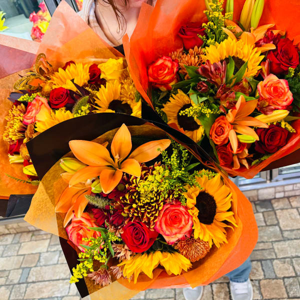 Three colorful bouquets with sunflowers, roses, and lilies wrapped in orange paper