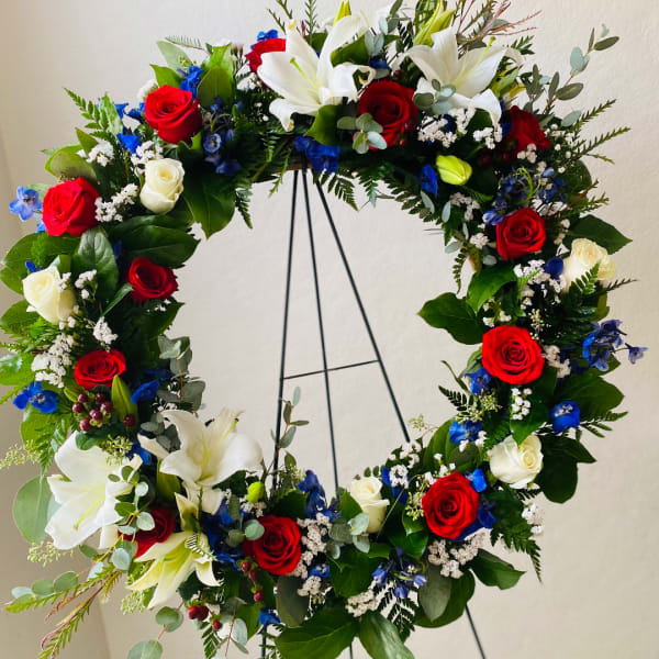 Circular floral wreath with red and white roses, white lilies, and blue accents