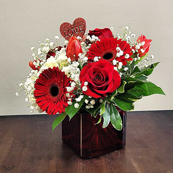 Red roses, gerbera daisies, and tulips with white accents in a red glass cube vase and heart LOVE pick