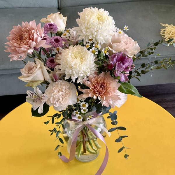 Vase of pastel pink, white, and purple flowers with a ribbon on a bright yellow table.