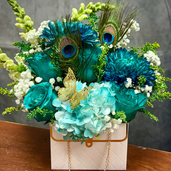 Teal and blue floral arrangement with peacock feathers in a white gift box.