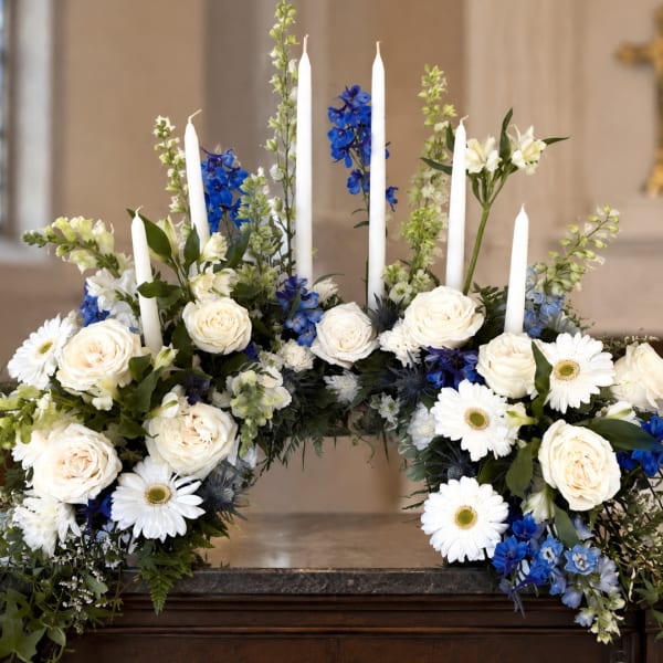White roses and daisies arranged with blue flowers and tall white candles
