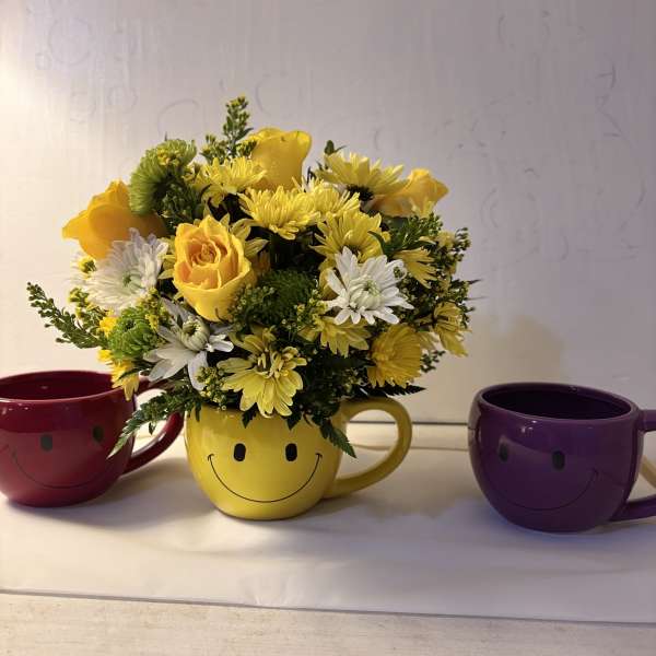 Yellow and white flower arrangement in a smiley-face mug, flanked by two colorful mugs