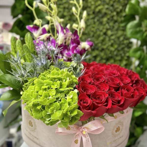 Round hatbox arrangement with red roses, green hydrangeas, and purple orchids tied with a pink ribbon.