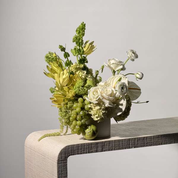 White and green floral arrangement in a vase with grapes and an apple