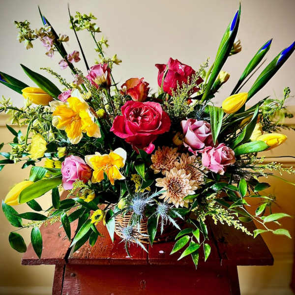 Mixed basket arrangement with yellow tulips and daffodils, red and pink roses, and mixed blooms on a wooden table.