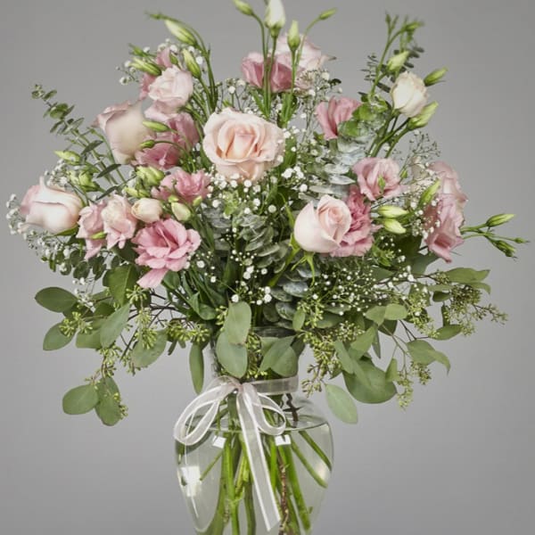 Tall arrangement of pale pink roses and lisianthus with baby's breath in a clear glass vase tied with a ribbon.