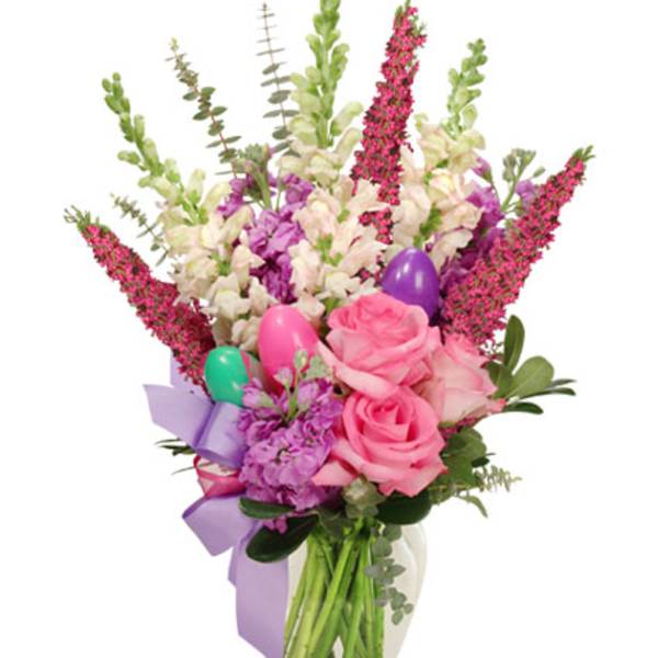 Tall pastel arrangement with pink roses, white snapdragons, purple blooms, and colorful eggs in a clear vase with a bow