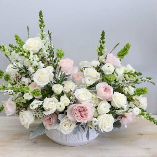 Low white bowl of white and blush roses with green accents in a wide, airy arrangement