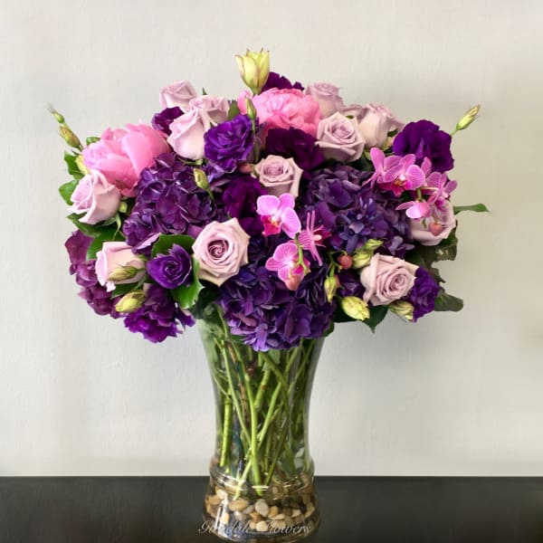 Tall glass vase of purple hydrangeas, lavender roses, and pink blooms in a rounded arrangement