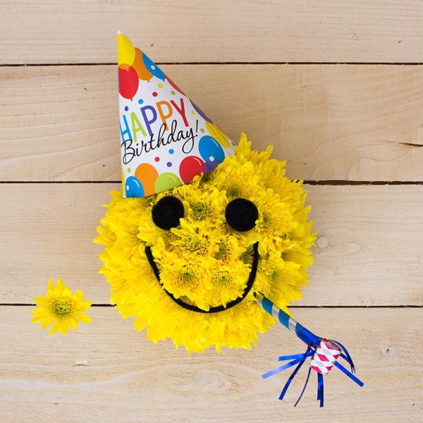 Round yellow mum arrangement shaped like a smiley face with birthday hat and party blower