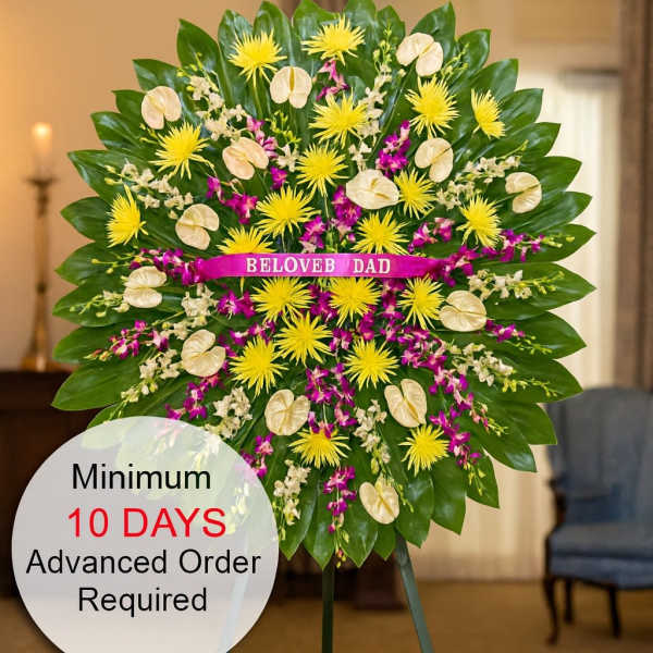 Tall standing sympathy spray with yellow mums, cream tropical blooms and purple orchids on a green leaf fan.