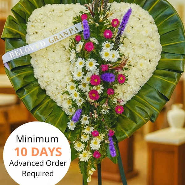 Heart-shaped white sympathy standing spray with cascading purple and pink flowers on a green-leaf border