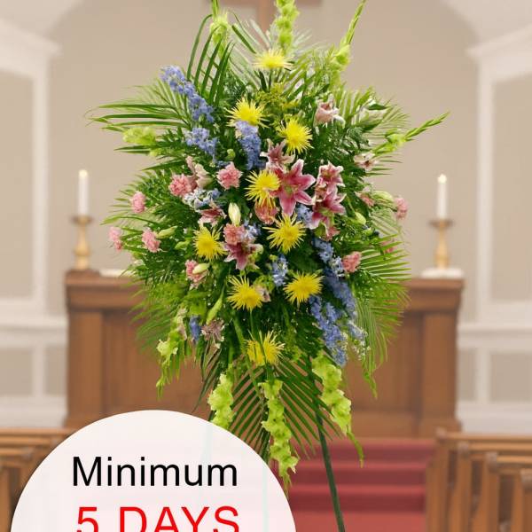 Tall standing funeral spray of pink, yellow, and blue flowers on an easel in a church aisle