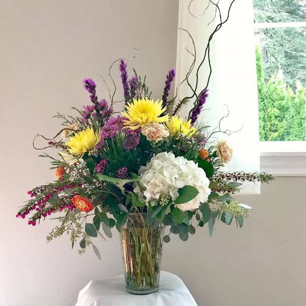 Tall mixed arrangement with yellow blooms, white hydrangea, and purple flowers in a clear glass vase.