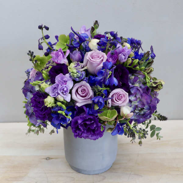 Round arrangement of purple, blue, and lavender flowers in a gray cylinder vase