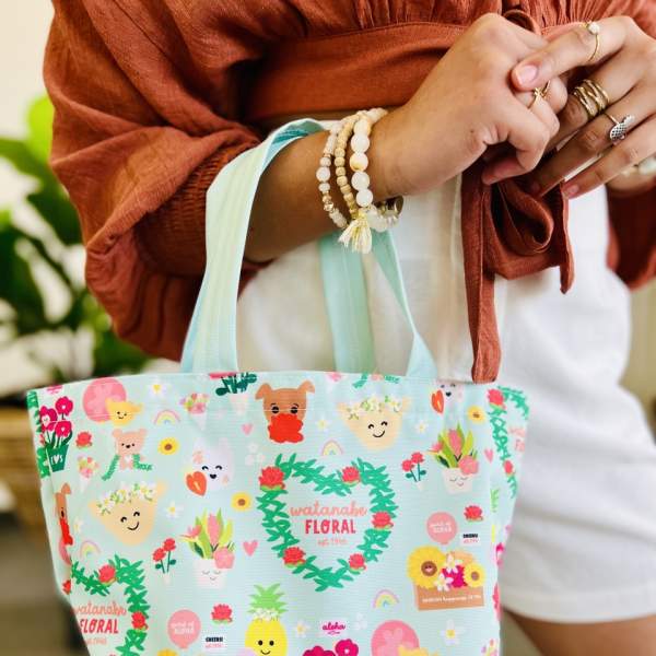 Small aqua tote bag with colorful cartoon floral and character print held by a person.