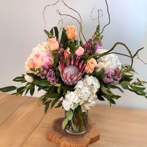Arrangement of protea, roses, hydrangeas and mixed blooms in a glass vase on a wooden slice