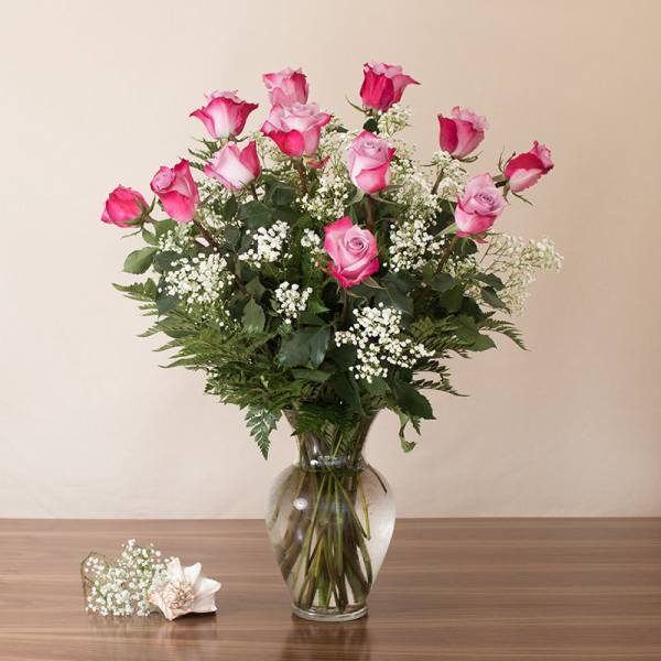 Tall arrangement of pink roses with white filler flowers in a clear glass vase on a wooden table