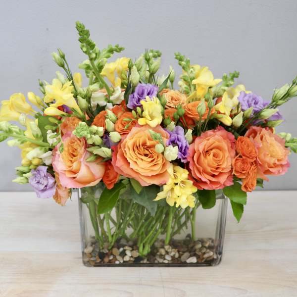 Low rectangular arrangement of orange roses, yellow flowers, and purple blooms in a glass vase with pebbles