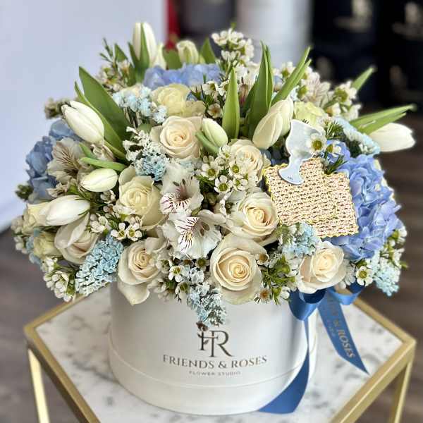 Round white hatbox of cream roses, white tulips, and blue hydrangeas with small white blossoms and a blue ribbon.