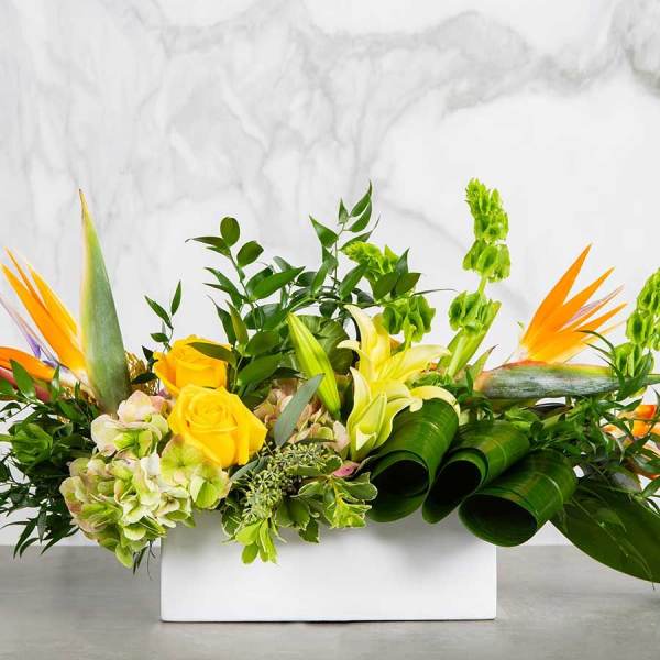 Low rectangular arrangement with yellow roses, lilies, and tropical flowers in a white container.
