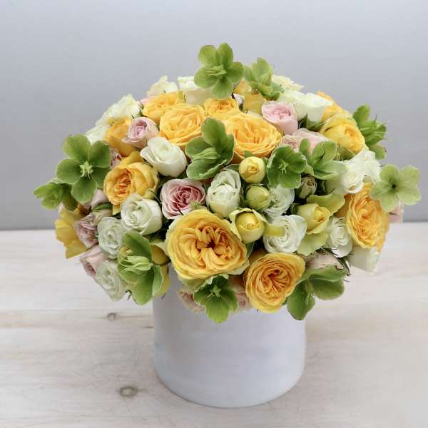Low arrangement of yellow and pastel roses with green blooms in a white ceramic cylinder vase