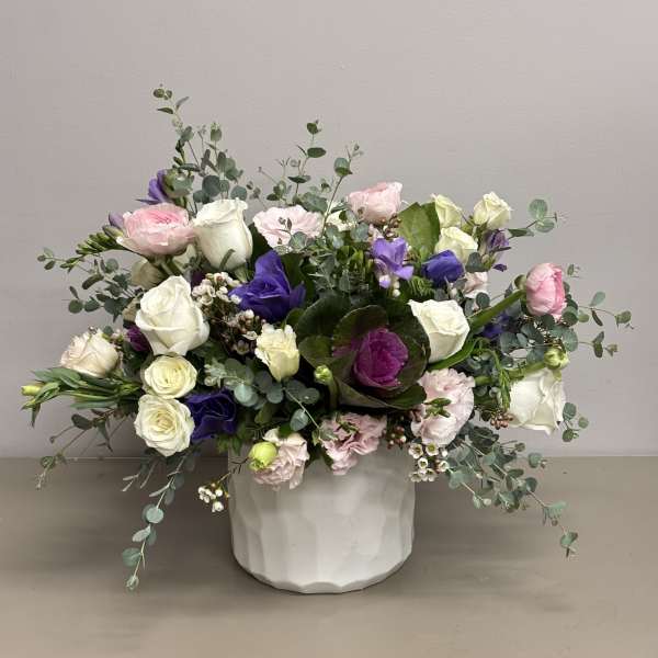 Low arrangement of white roses, pink blooms, and purple flowers in a white ceramic pot