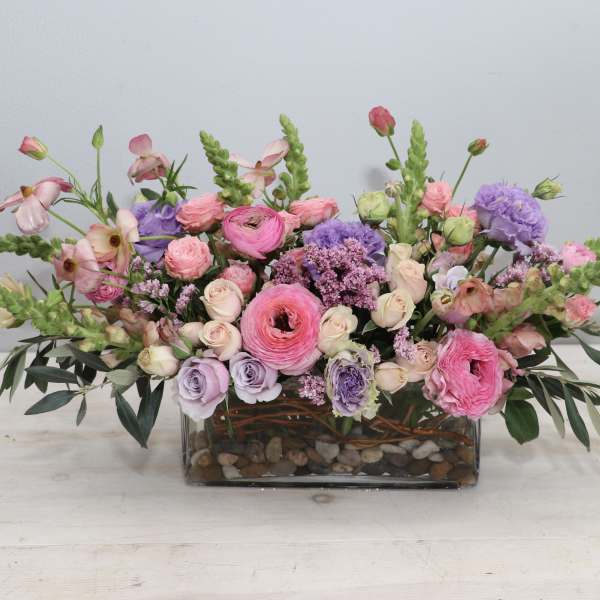 Low rectangular arrangement of pink and lavender flowers in a glass vase with river rocks
