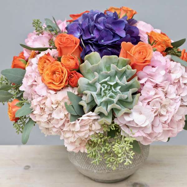 Low bowl arrangement of pink hydrangeas, orange roses, purple blooms, and a central green succulent