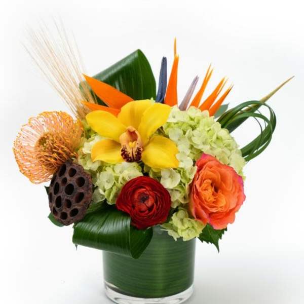 Colorful tropical arrangement with hydrangeas, orchid, rose, and protea in a leaf-wrapped glass vase