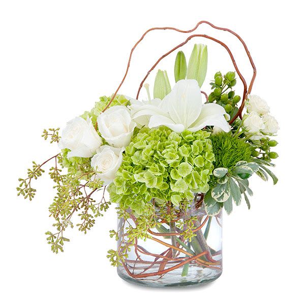 Low arrangement of white lilies, white roses, and green hydrangeas in a clear glass vase with curly branches.