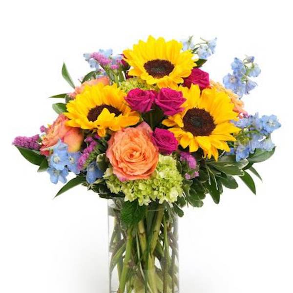 Arrangement of yellow sunflowers, pink roses, blue delphinium, and green hydrangeas in a clear glass vase.