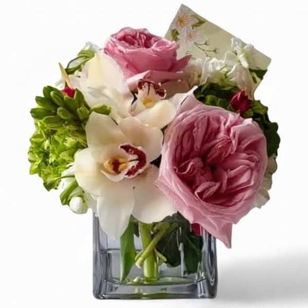 Compact arrangement of pink roses and white orchids in a clear square glass vase with a card.