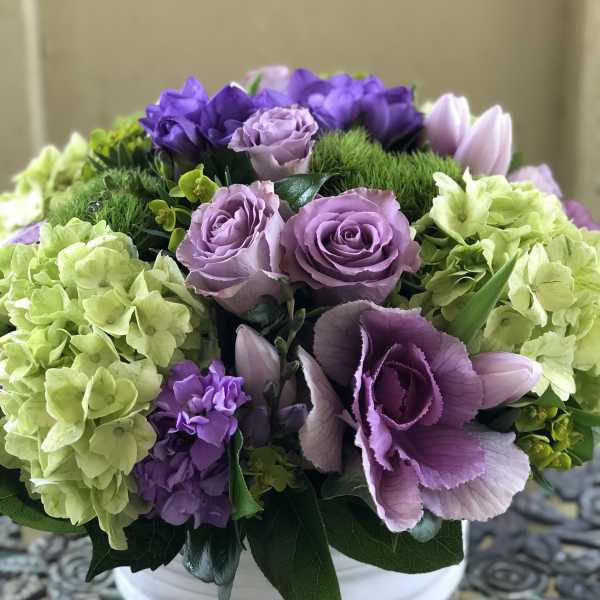 Low arrangement of lavender roses, green hydrangeas, and purple blooms in a white bowl