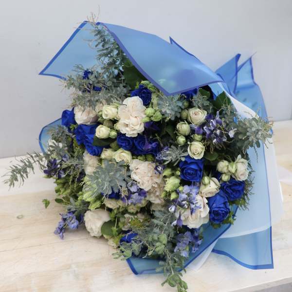 Bouquet of blue and cream roses with small white buds wrapped in blue cellophane.