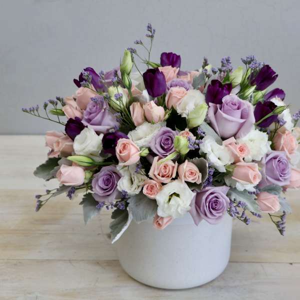Low pastel arrangement of pink and lavender roses with white blooms in a white ceramic pot