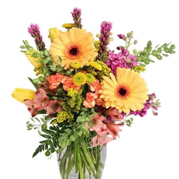 Mixed bouquet of yellow gerbera daisies, pink blooms, and purple stock in a clear glass vase