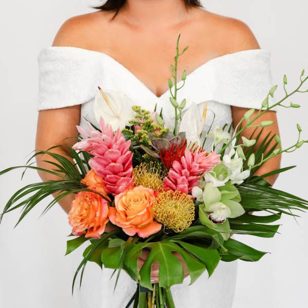 Bride in an off-shoulder white dress holding a large tropical bouquet of pink, orange, green, and white flowers.