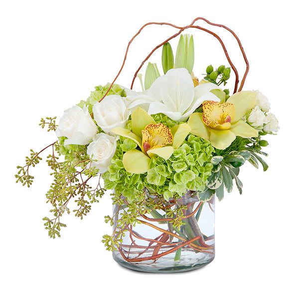 Low arrangement of white lilies and roses with green hydrangeas and yellow orchids in a clear glass vase.