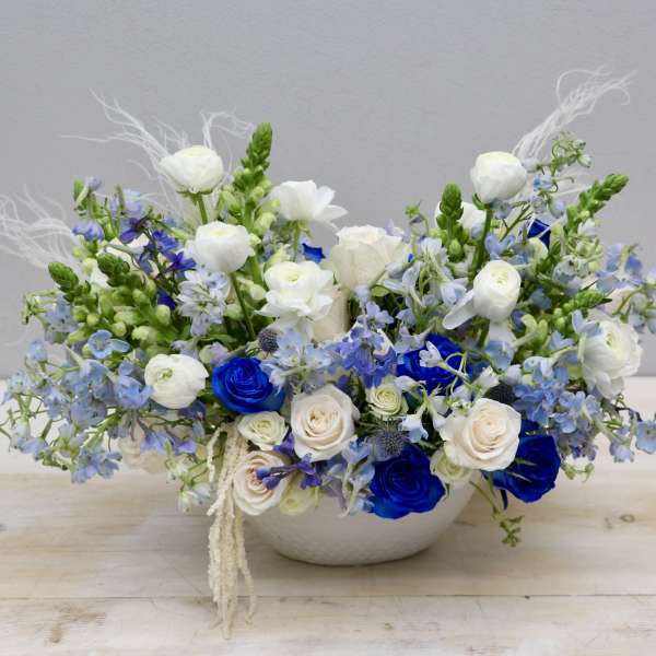 Low white bowl arrangement of blue and white roses with white ranunculus and light blue flowers
