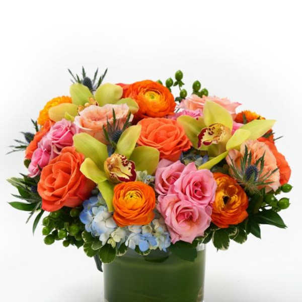 Low round arrangement of orange and pink roses, orchids, and ranunculus in a clear glass vase