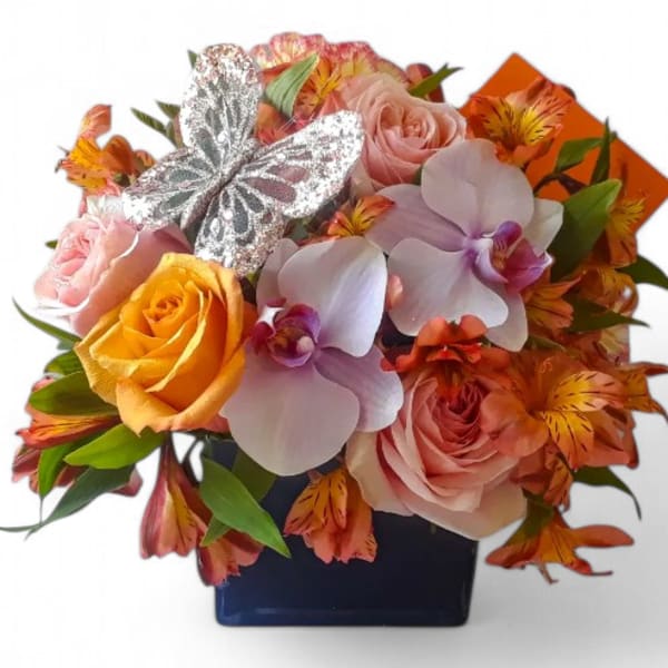 Compact arrangement of roses, orchids, and alstroemeria in a blue cube vase with a silver butterfly decoration