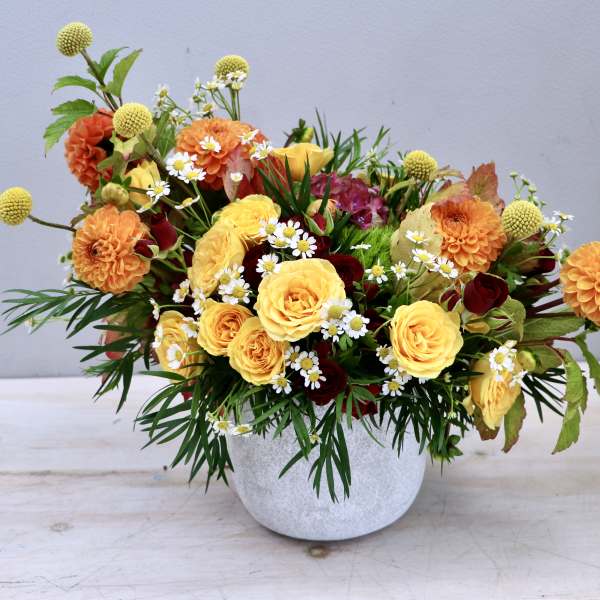 Low arrangement of yellow roses, orange dahlias, and small white daisies in a white pot
