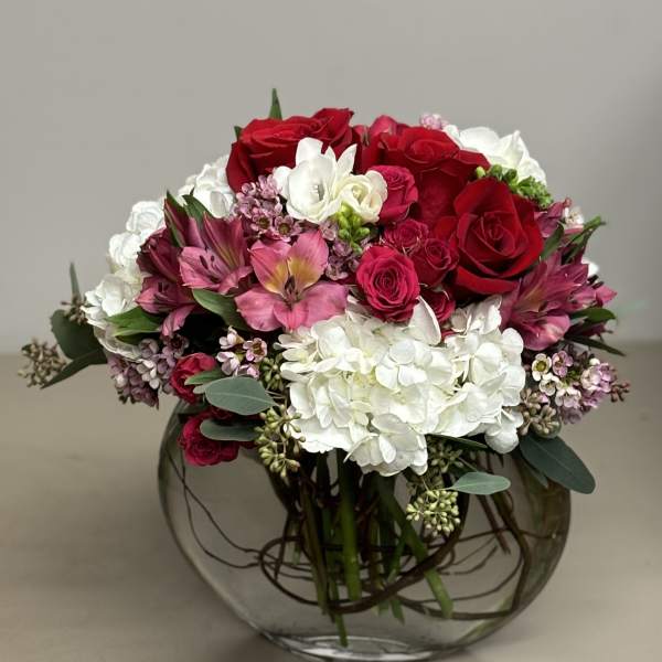 Low round arrangement of red roses, pink blooms, and white hydrangeas in a clear glass bowl vase