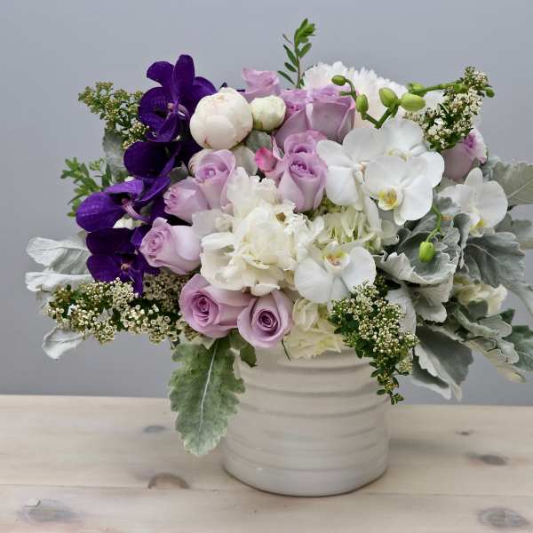 Low white vase filled with lavender roses, white hydrangeas, and orchids with purple accents