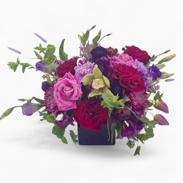 Low arrangement of red and pink roses with purple blooms and a green orchid in a black cube vase